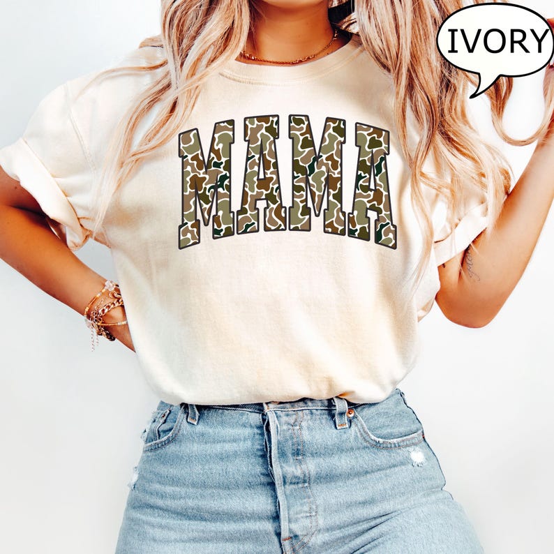 May include: A white t-shirt with the word "MAMA" printed in a camouflage pattern. The shirt is being worn by a person with long blonde hair and is available in the color ivory.