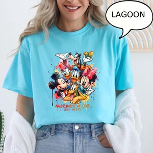 May include: A light blue t-shirt featuring a colorful graphic of Disney characters including Mickey Mouse, Minnie Mouse, Donald Duck, and Goofy. The graphic is accented with paint splatters and the text "Mickey & Co. EST 1928". A speech bubble says "LAGOON".