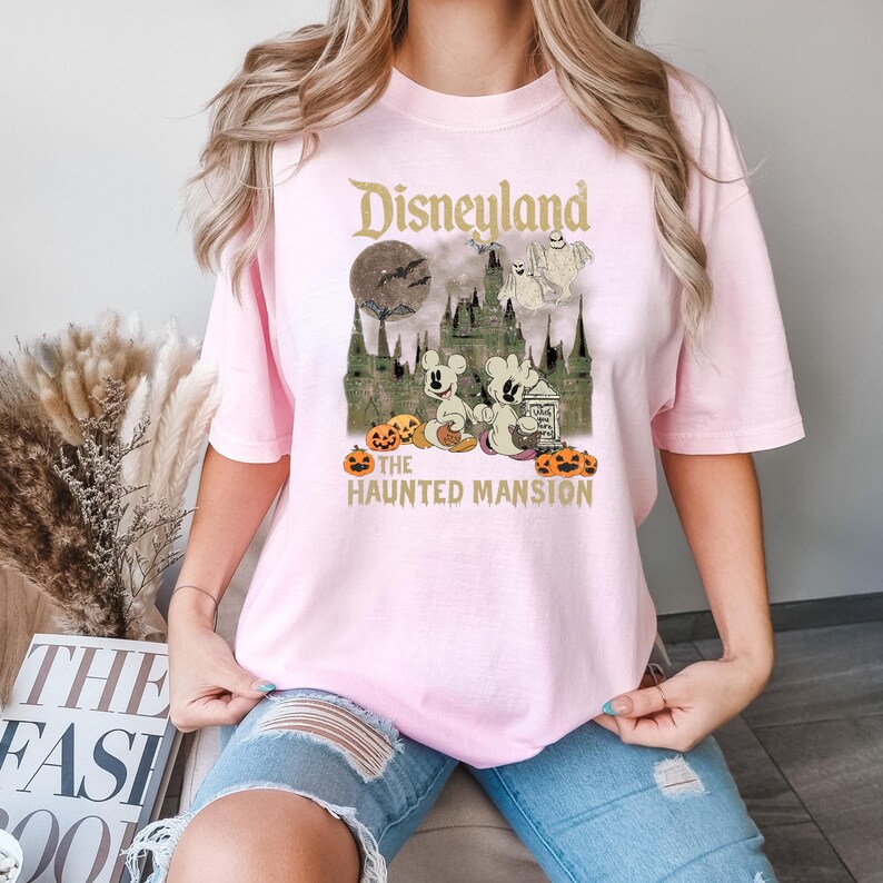 May include: Pink t-shirt with a vintage-style graphic of Mickey Mouse and friends at Disneyland's Haunted Mansion. The graphic features a spooky castle, pumpkins, and ghosts. The text "Disneyland" and "The Haunted Mansion" are printed on the shirt.