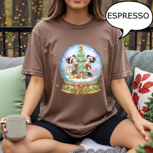 May include: Espresso-colored t-shirt featuring a snow globe design with Mickey and Minnie Mouse in festive attire. The snow globe includes a Christmas tree, gifts, and candy canes. The shirt has a crew neck and short sleeves.