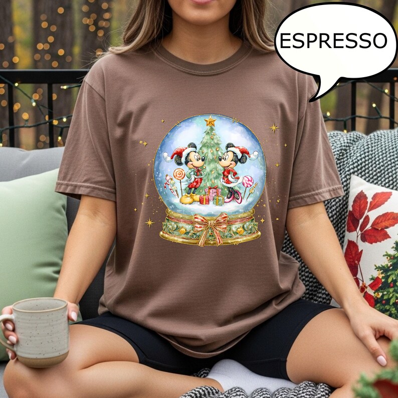 May include: Espresso-colored t-shirt featuring a snow globe design with Mickey and Minnie Mouse in festive attire. The snow globe includes a Christmas tree, gifts, and candy canes. The shirt has a crew neck and short sleeves.