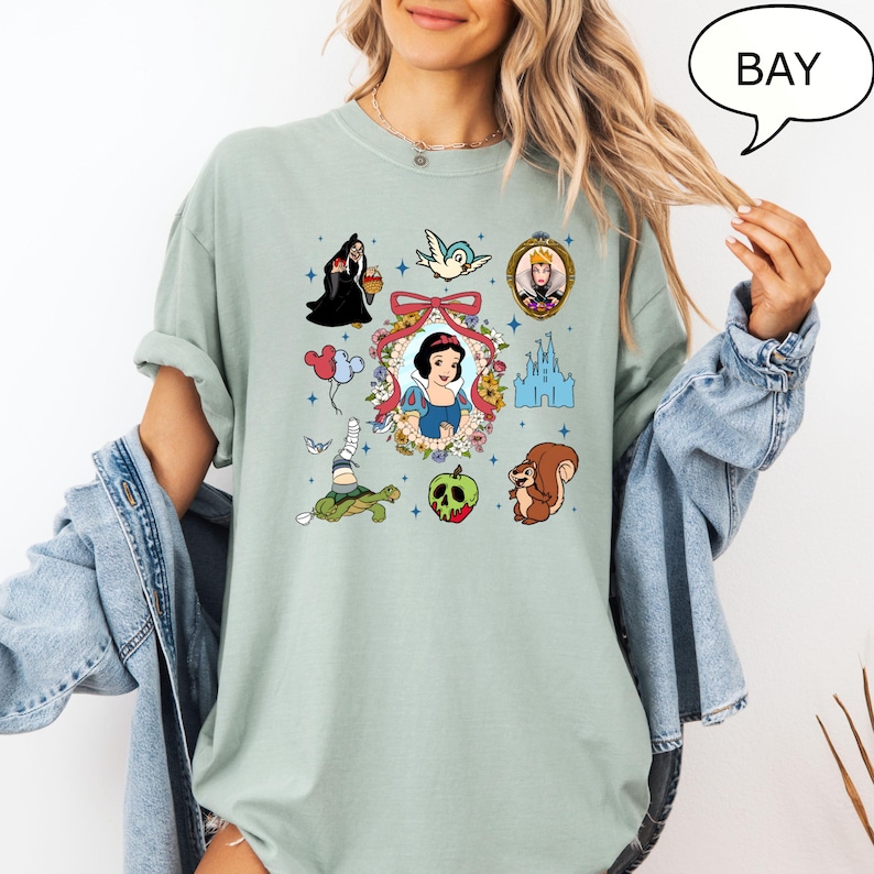 May include: A light green t-shirt with a graphic print of various Disney characters from Snow White and the Seven Dwarfs. The characters include Snow White, the Evil Queen, a bird, a squirrel, a turtle, and a castle. The text "BAY" is in a speech bubble in the upper right corner of the image.