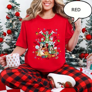 Puede incluir: Una camiseta roja con un gr&aacute;fico navide&ntilde;o de personajes de Disney, incluyendo Mickey y Minnie Mouse, Donald y Daisy Duck, y Goofy. El dise&ntilde;o incluye firmas y elementos festivos. La palabra "RED" est&aacute; en un bocadillo.
