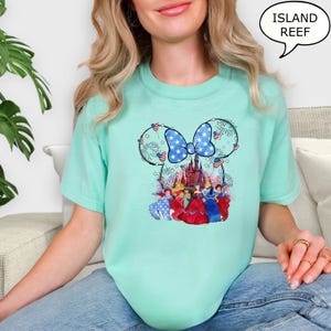 Puede incluir: Camiseta verde azulado claro con un dise&ntilde;o de Minnie Mouse con un lazo patri&oacute;tico y fuegos artificiales. El dise&ntilde;o incluye un castillo y varias princesas de Disney. El texto "ISLAND REEF" est&aacute; en una burbuja.