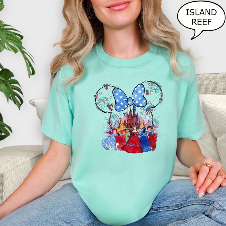 Puede incluir: Camiseta verde azulado claro con un dise&ntilde;o de Minnie Mouse con un lazo patri&oacute;tico y fuegos artificiales. El dise&ntilde;o incluye un castillo y varias princesas de Disney. El texto "ISLAND REEF" est&aacute; en una burbuja.