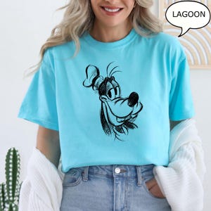 Puede incluir: Una camiseta azul claro con una ilustraci&oacute;n de dibujos animados en blanco y negro de Goofy. El texto "LAGOON" est&aacute; en un bocadillo de di&aacute;logo sobre la imagen.