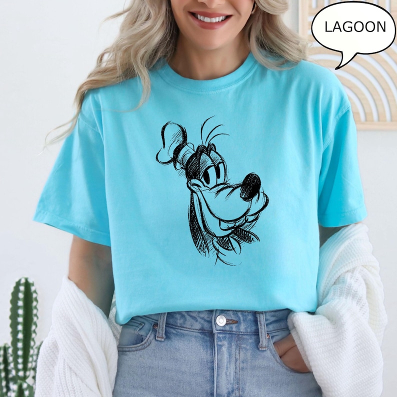 Puede incluir: Una camiseta azul claro con una ilustraci&oacute;n de dibujos animados en blanco y negro de Goofy. El texto "LAGOON" est&aacute; en un bocadillo de di&aacute;logo sobre la imagen.