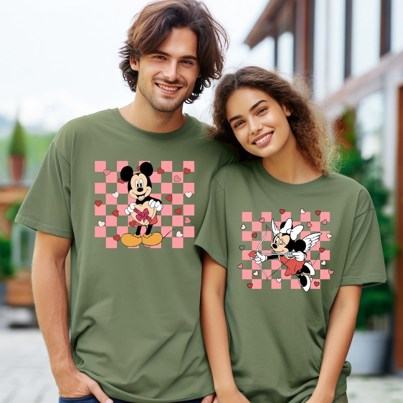 Puede incluir: Camisetas verde oliva con dise&ntilde;os de Mickey y Minnie Mouse. Mickey sostiene un regalo en forma de coraz&oacute;n sobre un fondo a cuadros rosa y blanco. Minnie est&aacute; vestida como Cupido con arco y flecha.
