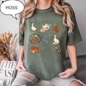 Puede incluir: Una camiseta de manga corta color verde musgo con un estampado gr&aacute;fico de varias ilustraciones enmarcadas que incluyen gansos, una tetera con taza, fresas en una cesta, bayas rojas en una rama y flores azules, con el texto MOSS en un globo.