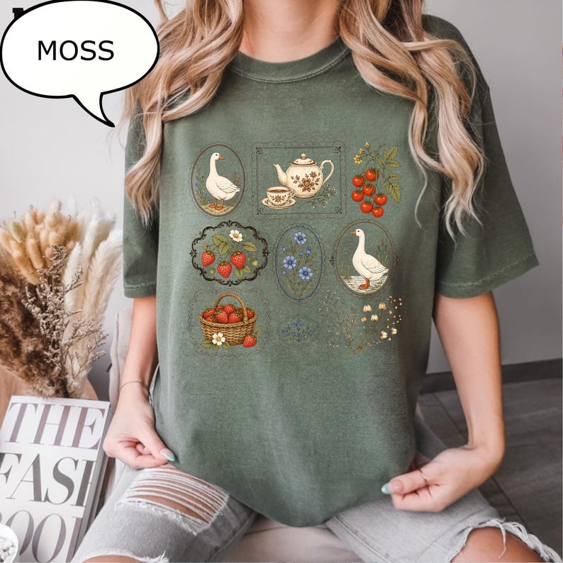 Puede incluir: Una camiseta de manga corta color verde musgo con un estampado gr&aacute;fico de varias ilustraciones enmarcadas que incluyen gansos, una tetera con taza, fresas en una cesta, bayas rojas en una rama y flores azules, con el texto MOSS en un globo.