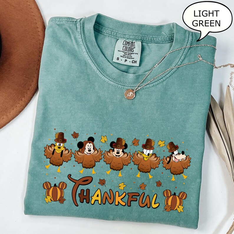 May include: Light green t-shirt with a Thanksgiving-themed graphic featuring cartoon characters dressed as turkeys. The word "THANKFUL" is printed in a decorative font. A silver necklace with a pendant is draped across the shirt.