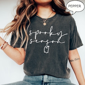 May include: Dark grey t-shirt featuring the words "spooky season" in white script, along with a ghost graphic. The shirt has a relaxed fit and is made of a soft material. The word "pepper" is in a speech bubble.