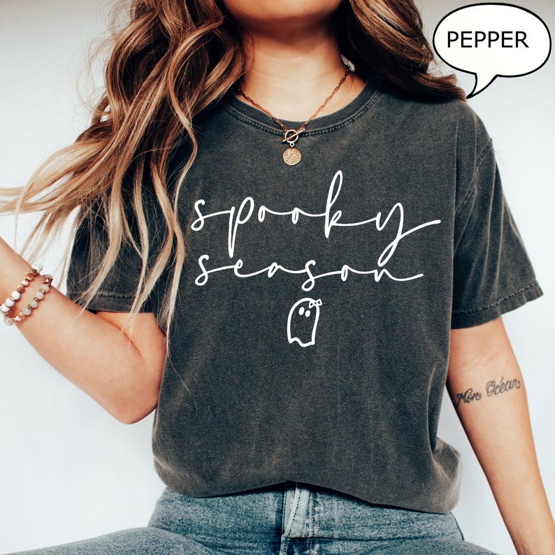 May include: Dark grey t-shirt featuring the words "spooky season" in white script, along with a ghost graphic. The shirt has a relaxed fit and is made of a soft material. The word "pepper" is in a speech bubble.