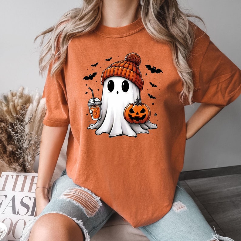 May include: Orange t-shirt featuring a cartoon ghost wearing a red and white knitted hat, holding a pumpkin and a cup of orange juice. The ghost has black eyes and a small mouth. There are bats and stars around the ghost.