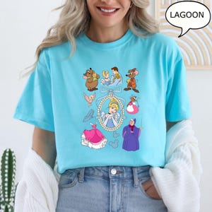 Puede incluir: Camiseta azul claro con ilustraciones de dibujos animados de un cuento de hadas. El dise&ntilde;o incluye una princesa, ratones, un hada madrina y otros personajes. La palabra "LAGOON" est&aacute; en una burbuja.