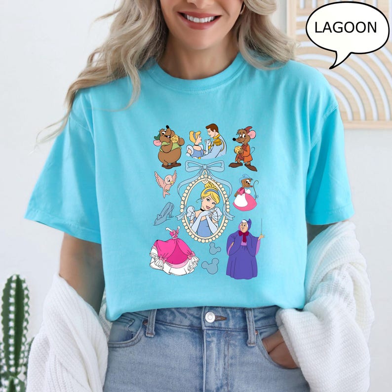 Puede incluir: Camiseta azul claro con ilustraciones de dibujos animados de un cuento de hadas. El dise&ntilde;o incluye una princesa, ratones, un hada madrina y otros personajes. La palabra "LAGOON" est&aacute; en una burbuja.