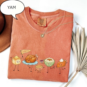 Pu&ograve; includere: Una t-shirt color terracotta con una grafica di cibi del Ringraziamento antropomorfi, tra cui un panino con una bandierina con la scritta THANKFUL, un tacchino arrosto, una ciotola di ripieno, pur&egrave; di patate e una fetta di torta.