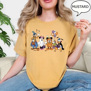 May include: Mustard-colored t-shirt featuring a Halloween-themed graphic of Disney characters in costumes. Goofy is dressed as a wizard, Minnie Mouse as a ghost, Mickey Mouse as a pumpkin, and Donald Duck as a vampire. The text "MUSTARD" is in a speech bubble.