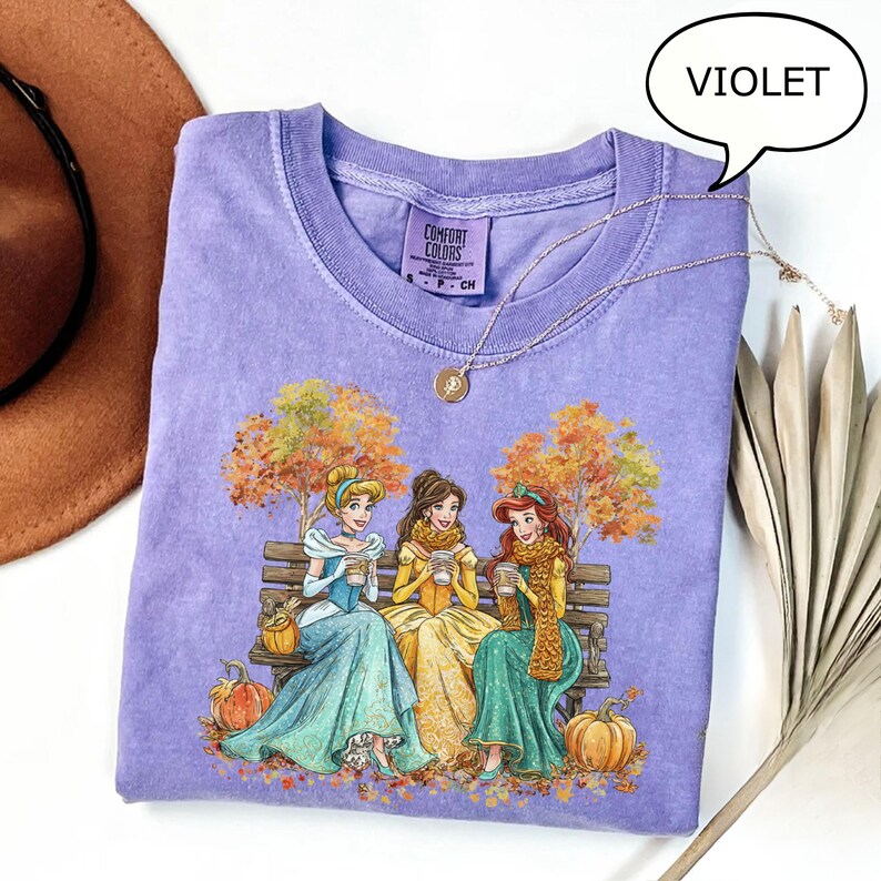 May include: Violet Comfort Colors t-shirt featuring a graphic of three princesses sitting on a bench, holding cups, with autumn trees and pumpkins. A gold necklace is draped over the shirt. The shirt is a light purple color.