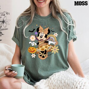 Puede incluir: Camiseta verde musgo con un dise&ntilde;o de Minnie Mouse de Halloween. Minnie est&aacute; disfrazada, sosteniendo una linterna de calabaza, con murci&eacute;lagos y flores. La palabra "MOSS" est&aacute; impresa en negro en la parte superior derecha.