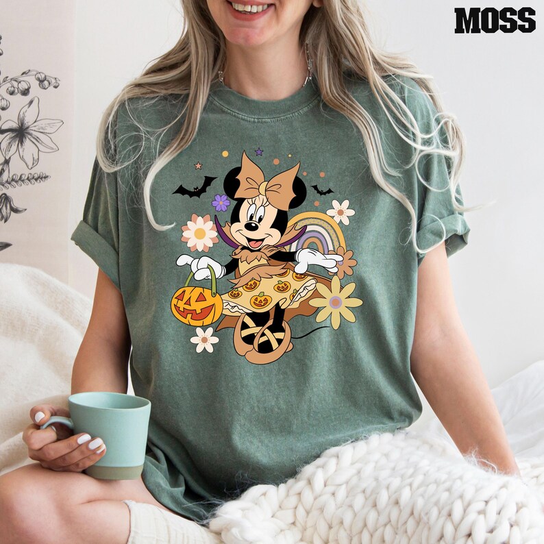 Puede incluir: Camiseta verde musgo con un dise&ntilde;o de Minnie Mouse de Halloween. Minnie est&aacute; disfrazada, sosteniendo una linterna de calabaza, con murci&eacute;lagos y flores. La palabra "MOSS" est&aacute; impresa en negro en la parte superior derecha.
