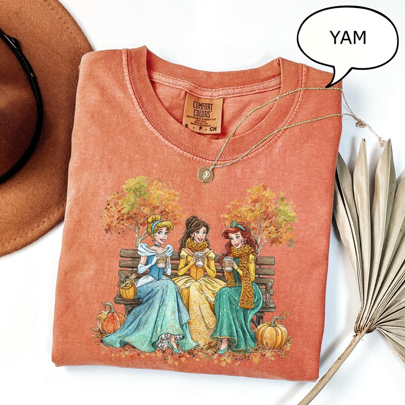 May include: Orange Comfort Colors t-shirt featuring a graphic of three cartoon princesses sitting on a bench, holding cups, with fall foliage and pumpkins. A gold necklace and the word "YAM" in a speech bubble are also visible.