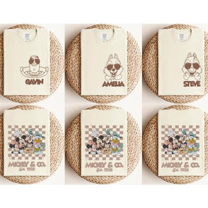 May include: Cream-colored t-shirts featuring cartoon characters with sunglasses and names like "GAVIN", "AMELIA", and "STEVE". Other shirts display Mickey Mouse and friends with "MICKEY & CO. Est. 1928" on a checkered background. The shirts are displayed on woven mats.