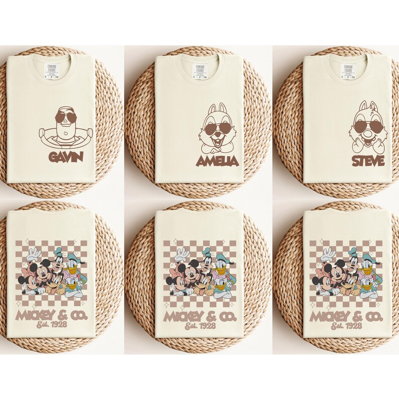 May include: Cream-colored t-shirts featuring cartoon characters with sunglasses and names like "GAVIN", "AMELIA", and "STEVE". Other shirts display Mickey Mouse and friends with "MICKEY & CO. Est. 1928" on a checkered background. The shirts are displayed on woven mats.