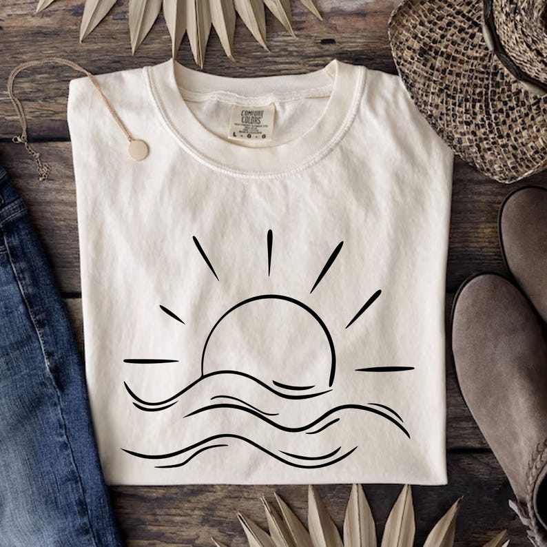 May include: A white t-shirt with a black line drawing of a sun setting over waves. The shirt is laid flat on a wooden surface.