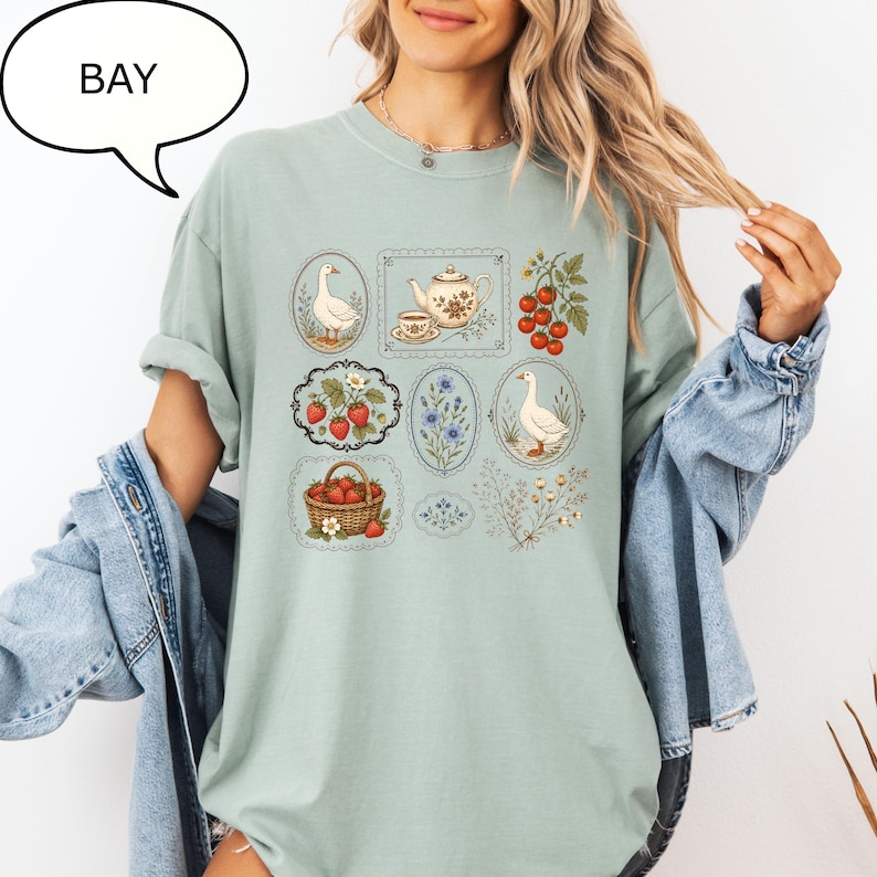 Puede incluir: Una camiseta verde salvia con un estampado gr&aacute;fico de varias ilustraciones que incluyen gansos, una tetera con taza, fresas en una cesta, tomates en rama y ramitas de flores. La palabra BAY est&aacute; impresa en un bocadillo sobre la camiseta.