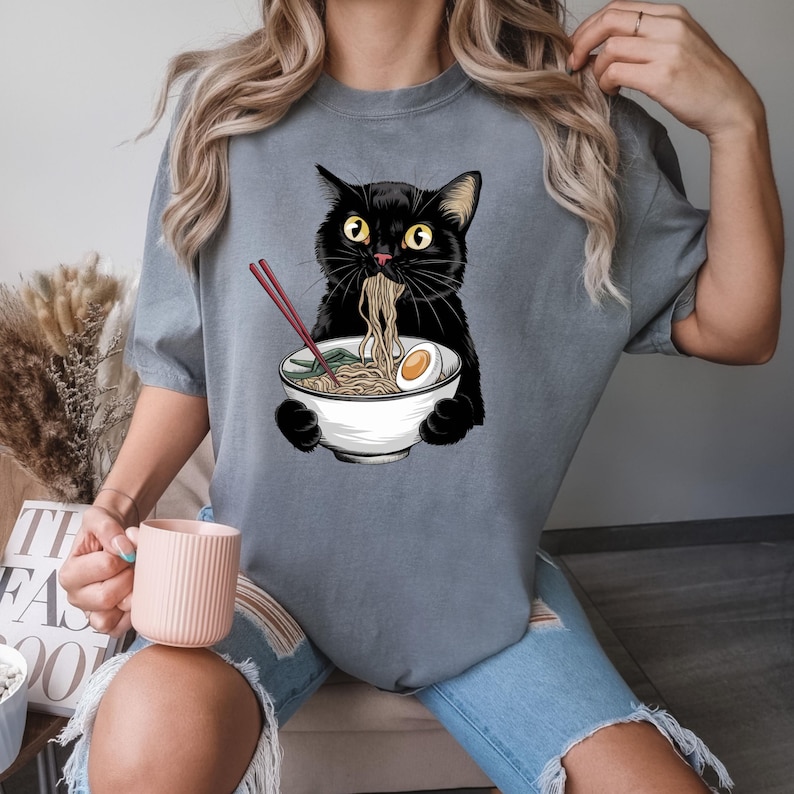 Puede incluir: Una camiseta gris con un gato negro comiendo fideos ramen de un taz&oacute;n blanco. El gato tiene una expresi&oacute;n de sorpresa en su rostro.