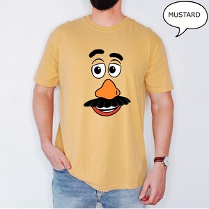 May include: Mustard-colored t-shirt featuring a cartoon face with a large orange nose, black mustache, and smiling red mouth. The eyes are large and white with black pupils. The word "MUSTARD" is in a speech bubble.