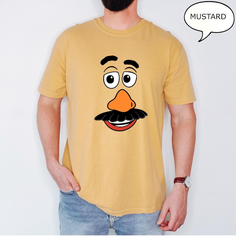 May include: Mustard-colored t-shirt featuring a cartoon face with a large orange nose, black mustache, and smiling red mouth. The eyes are large and white with black pupils. The word "MUSTARD" is in a speech bubble.