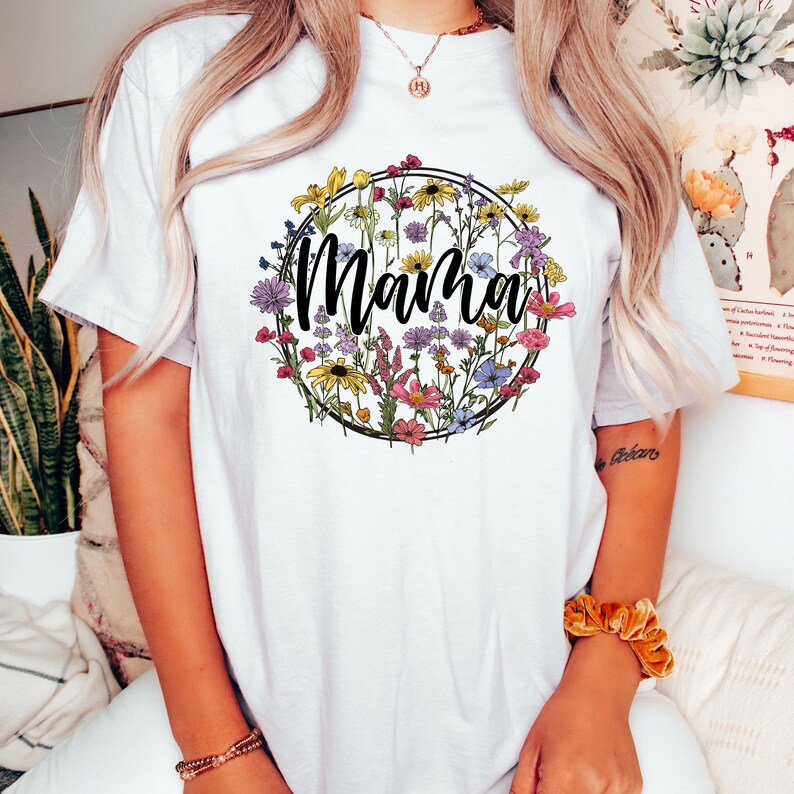 Peut inclure: T-shirt blanc avec un motif circulaire floral et le mot "Mama" en lettres noires.