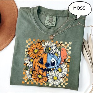May include: Green Comfort Colors t-shirt with a graphic featuring a pumpkin and a blue character surrounded by orange and white flowers. The shirt has a small tag and a silver necklace. The word "MOSS" is in a speech bubble.