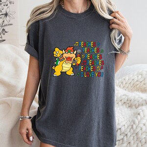 May include: Dark gray t-shirt featuring a cartoon Bowser character holding a microphone. The text "PEACHES I LOVE YOU" is printed in colorful letters with musical notes. The shirt is a comfortable, casual style.