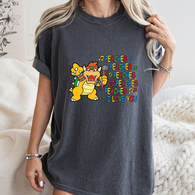 May include: Dark gray t-shirt featuring a cartoon Bowser character holding a microphone. The text "PEACHES I LOVE YOU" is printed in colorful letters with musical notes. The shirt is a comfortable, casual style.