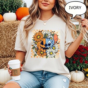 May include: Ivory-colored t-shirt featuring a Halloween-themed graphic. The design includes a jack-o'-lantern, a blue character, orange and white flowers, and bats. The shirt is a relaxed fit and suitable for casual wear.