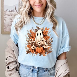 Op de afbeelding: Een lichtblauw T-shirt met een witte geest omringd door oranje en witte bloemen en een enkele oranje pompoen. De geest heeft een vriendelijk gezicht en is omringd door herfstbladeren.
