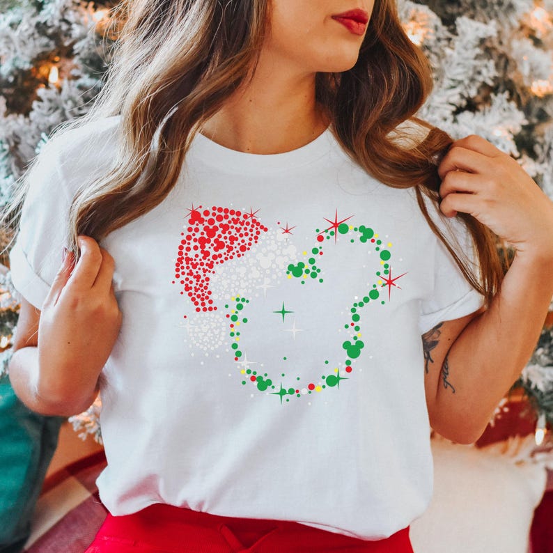 Puede incluir: Camiseta blanca con un dise&ntilde;o festivo. El dise&ntilde;o incluye un gorro de Pap&aacute; Noel y la forma de la cabeza de Mickey Mouse, creados con puntos y estrellas rojas, blancas, verdes y amarillas. Es una prenda informal con tem&aacute;tica navide&ntilde;a.