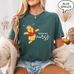 May include: A blue spruce t-shirt featuring a cartoon bear in a red jacket and nurse's hat, with the text "Pediatric Nurse". The design includes bees. The shirt is a comfortable, casual style.