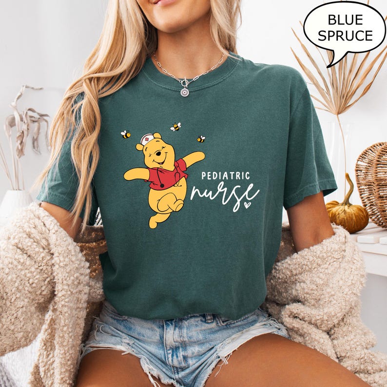 May include: A blue spruce t-shirt featuring a cartoon bear in a red jacket and nurse's hat, with the text "Pediatric Nurse". The design includes bees. The shirt is a comfortable, casual style.