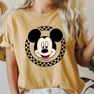 Puede incluir: Una camiseta amarilla con un c&iacute;rculo a cuadros blanco y negro con una imagen de dibujos animados de la cara de Mickey Mouse.