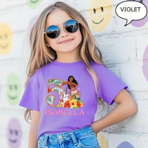 May include: A violet t-shirt featuring a colorful graphic of a cartoon character, the number 5, and the name "ISABELLA". The graphic includes tropical flowers and foliage. The girl in the image is wearing sunglasses.