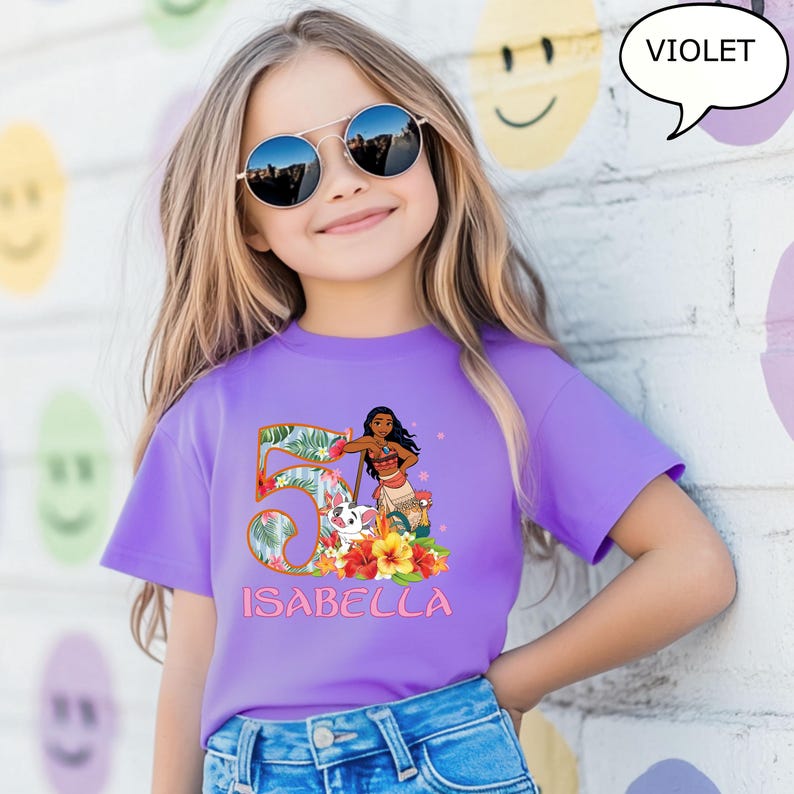 May include: A violet t-shirt featuring a colorful graphic of a cartoon character, the number 5, and the name "ISABELLA". The graphic includes tropical flowers and foliage. The girl in the image is wearing sunglasses.