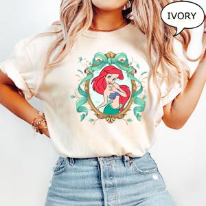 May include: Ivory-colored t-shirt featuring a framed illustration of Ariel from The Little Mermaid. The artwork showcases Ariel with red hair, a blue seashell bra, and a green tail, surrounded by a teal ribbon and floral accents. The word "IVORY" is in a speech bubble.