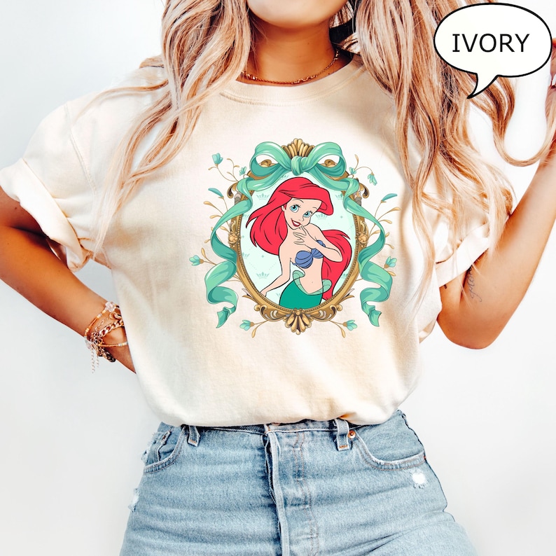 May include: Ivory-colored t-shirt featuring a framed illustration of Ariel from The Little Mermaid. The artwork showcases Ariel with red hair, a blue seashell bra, and a green tail, surrounded by a teal ribbon and floral accents. The word "IVORY" is in a speech bubble.