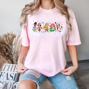 Puede incluir: Una camiseta rosa con un gr&aacute;fico de seis princesas Disney vestidas con atuendos navide&ntilde;os y sosteniendo adornos. Las princesas est&aacute;n de pie en una l&iacute;nea con luces navide&ntilde;as y copos de nieve a su alrededor.
