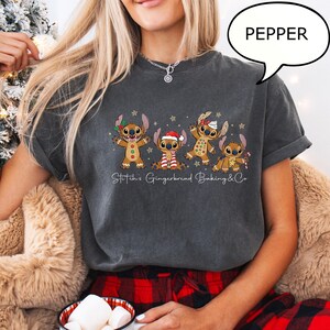 Puede incluir: Camiseta gris oscuro con un dise&ntilde;o de cuatro personajes Stitch con tem&aacute;tica de pan de jengibre y adornos navide&ntilde;os. El texto "Stitch's Gingerbread Baking & Co" est&aacute; impreso debajo. La camiseta tiene mangas cortas y un corte holgado.