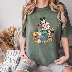 May include: A green t-shirt featuring a vintage-style illustration of Mickey Mouse, Minnie Mouse, and Pluto delivering presents. The illustration includes a mailbox with the word "Mail" on it and stars in the background.